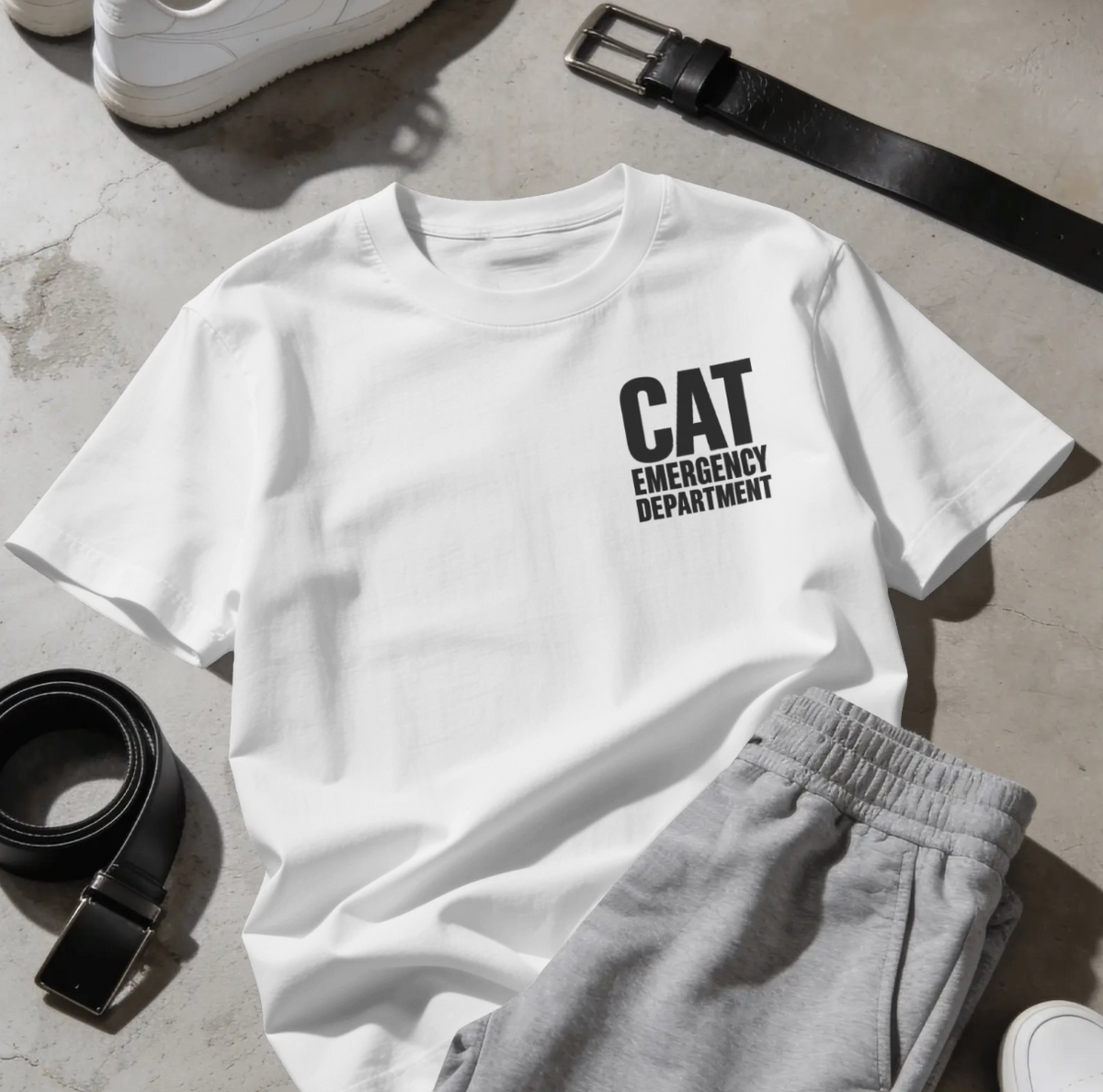 A white short-sleeve T-shirt laid flat on a light stone surface, printed with the words 'CAT EMERGENCY DEPARTMENT' in black text on the chest. Gray sweatpants, a black belt, and white sneakers are styled around it as outfit props.