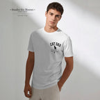 A man wearing a white short-sleeve crewneck t-shirt with a black 'CAT DAD' graphic on the chest, paired with light beige trousers, set against a minimalist light gray background with soft shadows.