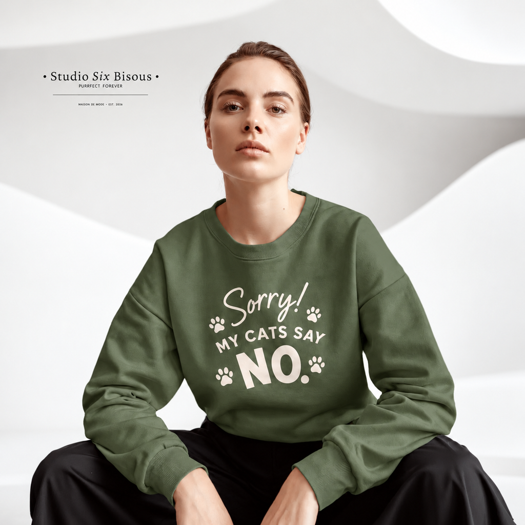 A woman wearing an olive green crewneck sweatshirt with the white printed text 'Sorry! MY CATS SAY NO.' and paw prints, seated against a white wavy studio background.
