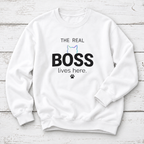 A white crewneck sweatshirt with a graphic print that reads 'THE REAL BOSS lives here.' in black text, featuring a small pastel cat outline and paw print, displayed flat on a white wood plank background.