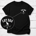 A black short-sleeve t-shirt with a white 'CAT DAD' graphic on the left chest, displayed on a white wood plank background with a circular zoom-in detail overlay.