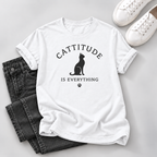 A white short-sleeve graphic T-shirt laid flat on a light gray surface, styled with folded dark gray jeans, white sneakers, and a small black paw-print detail. The shirt graphic reads “CATTITUDE IS EVERYTHING” with a black cat silhouette.
