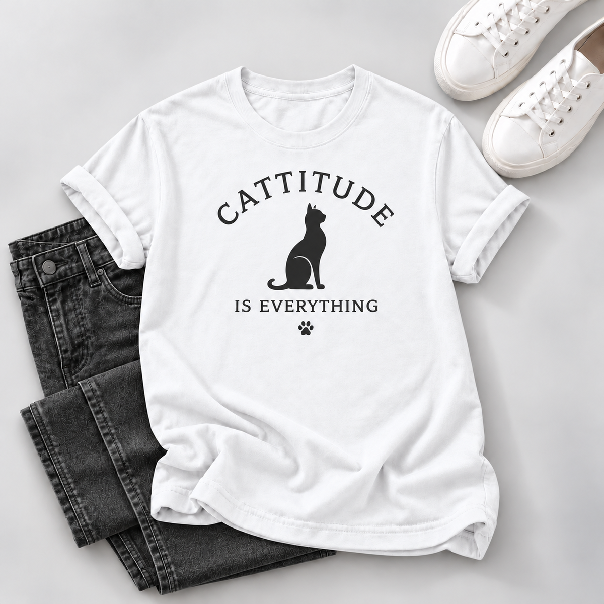 A white short-sleeve graphic T-shirt laid flat on a light gray surface, styled with folded dark gray jeans, white sneakers, and a small black paw-print detail. The shirt graphic reads “CATTITUDE IS EVERYTHING” with a black cat silhouette.