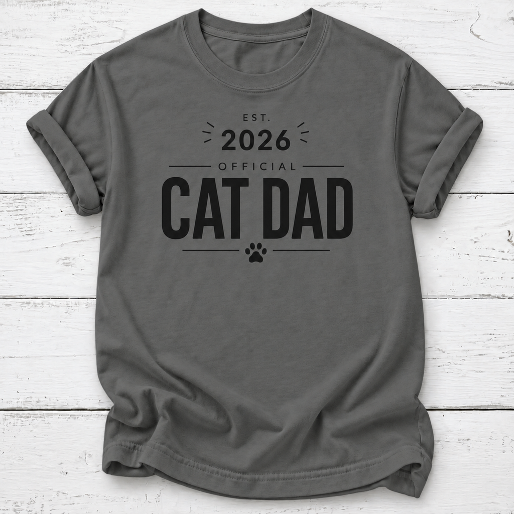 A dark gray crewneck t-shirt with a black graphic print that reads 'EST. 2026 OFFICIAL CAT DAD' with a paw print, laid flat on a white wood plank background.