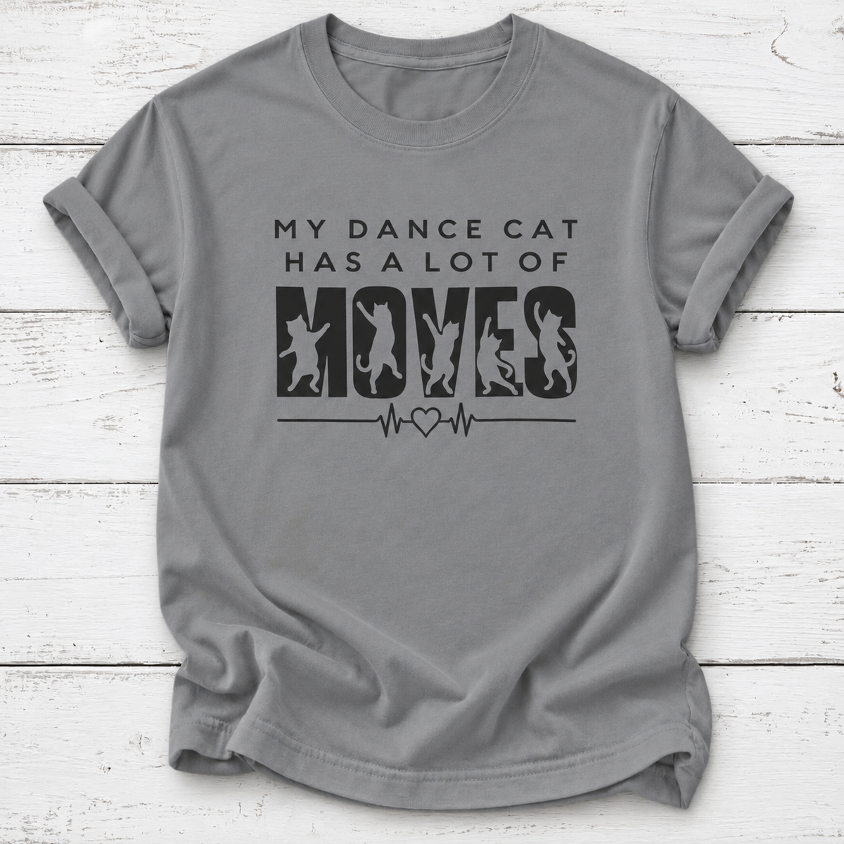 A gray short-sleeve crewneck t-shirt with a black graphic print that reads 'MY DANCE CAT HAS A LOT OF MOVES' with cat silhouettes inside the letters 'M-O-V-E-S' and a heartbeat line. The t-shirt is laid flat on a white wood plank background.