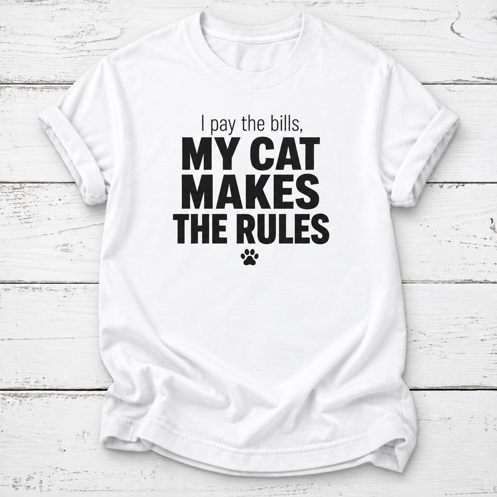 White T-shirt with black “I pay the bills, my cat makes the rules” lettering and a paw print, laid flat on white wooden boards.