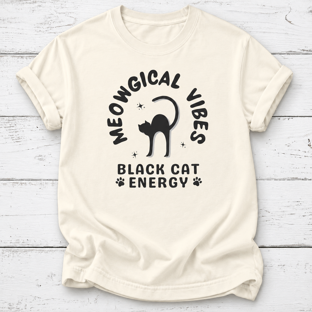 An ivory short-sleeve t-shirt with a black graphic print that reads 'MEOWGICAL VIBES BLACK CAT ENERGY' with a cat silhouette, laid flat on a white wood plank background.