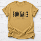 A mustard yellow short-sleeve t-shirt with black printed text reading 'MY CAT HAS BETTER BOUNDARIES THAN I DO.' laid flat on a white wood plank background.
