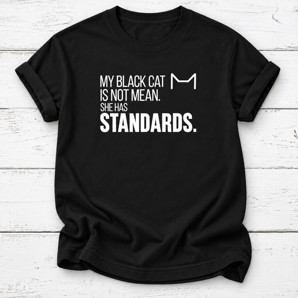 Black T-shirt with white “My Black Cat Is Not Mean. She Has Standards.” lettering and a minimalist cat-ear outline, laid flat on white wooden boards.