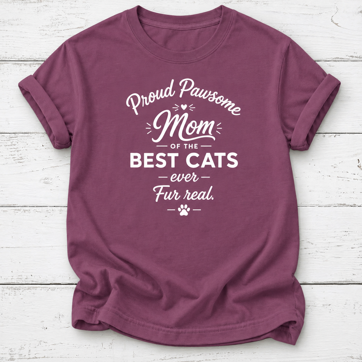 A mauve short-sleeve t-shirt with a white graphic text print reading 'Proud Pawsome Mom of the BEST CATS ever Fur real.' with a paw print, laid flat on a white wood plank background.