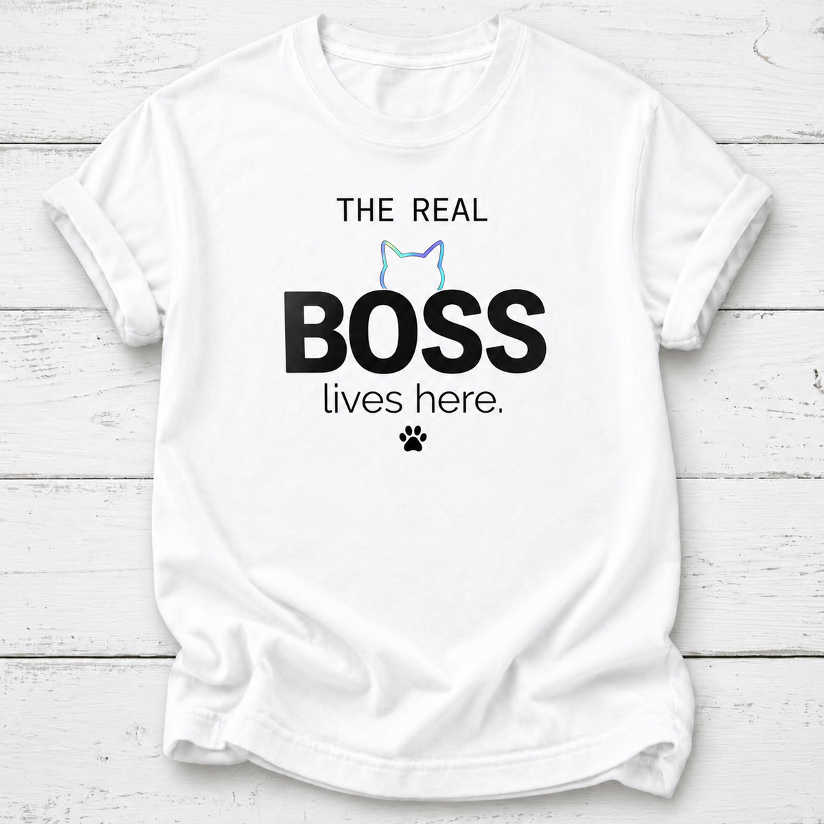 A white short-sleeve crewneck t-shirt laid flat on a white wood plank background. The shirt features a graphic print that reads 'THE REAL BOSS lives here.