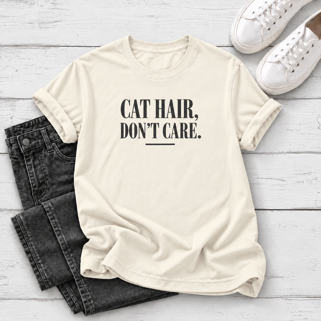 An off-white short-sleeve t-shirt with the text 'CAT HAIR, DON'T CARE.' printed in black on the front, styled in a flat lay with dark gray jeans and white sneakers on a white wood background.