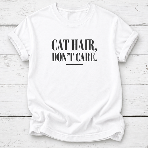 A white short-sleeve crewneck t-shirt with the text 'CAT HAIR, DON'T CARE.' printed in black serif font on the chest, laid flat on a white wood plank background.
