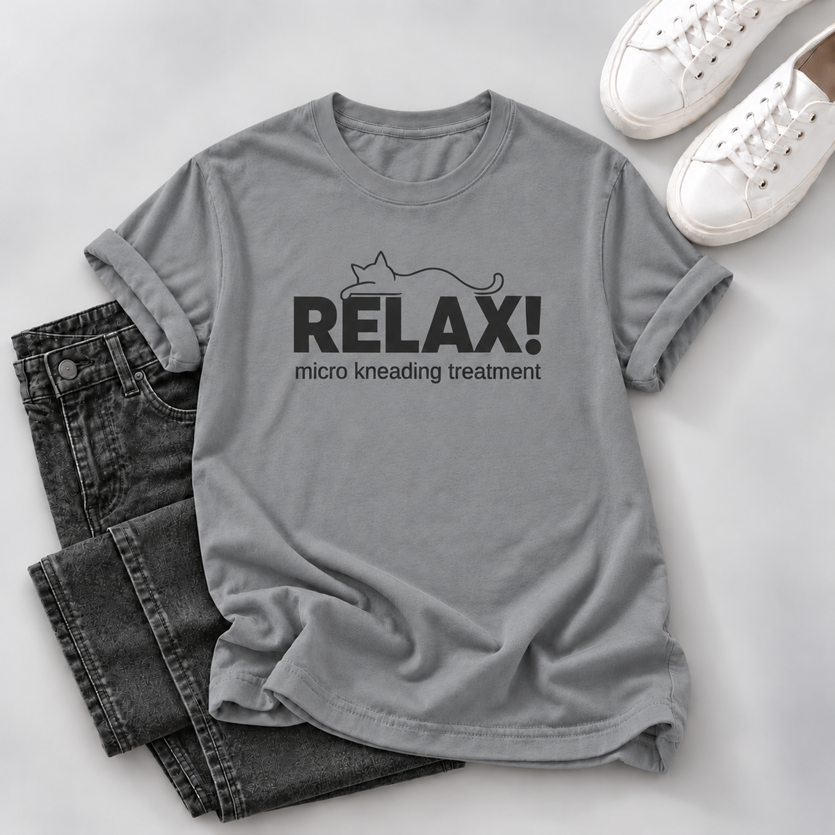 A gray short-sleeve T-shirt laid flat with rolled cuffs, featuring a black cat outline and the text 'RELAX! micro kneading treatment' on the front. The shirt is styled with folded dark jeans and white sneakers on a light gray background.