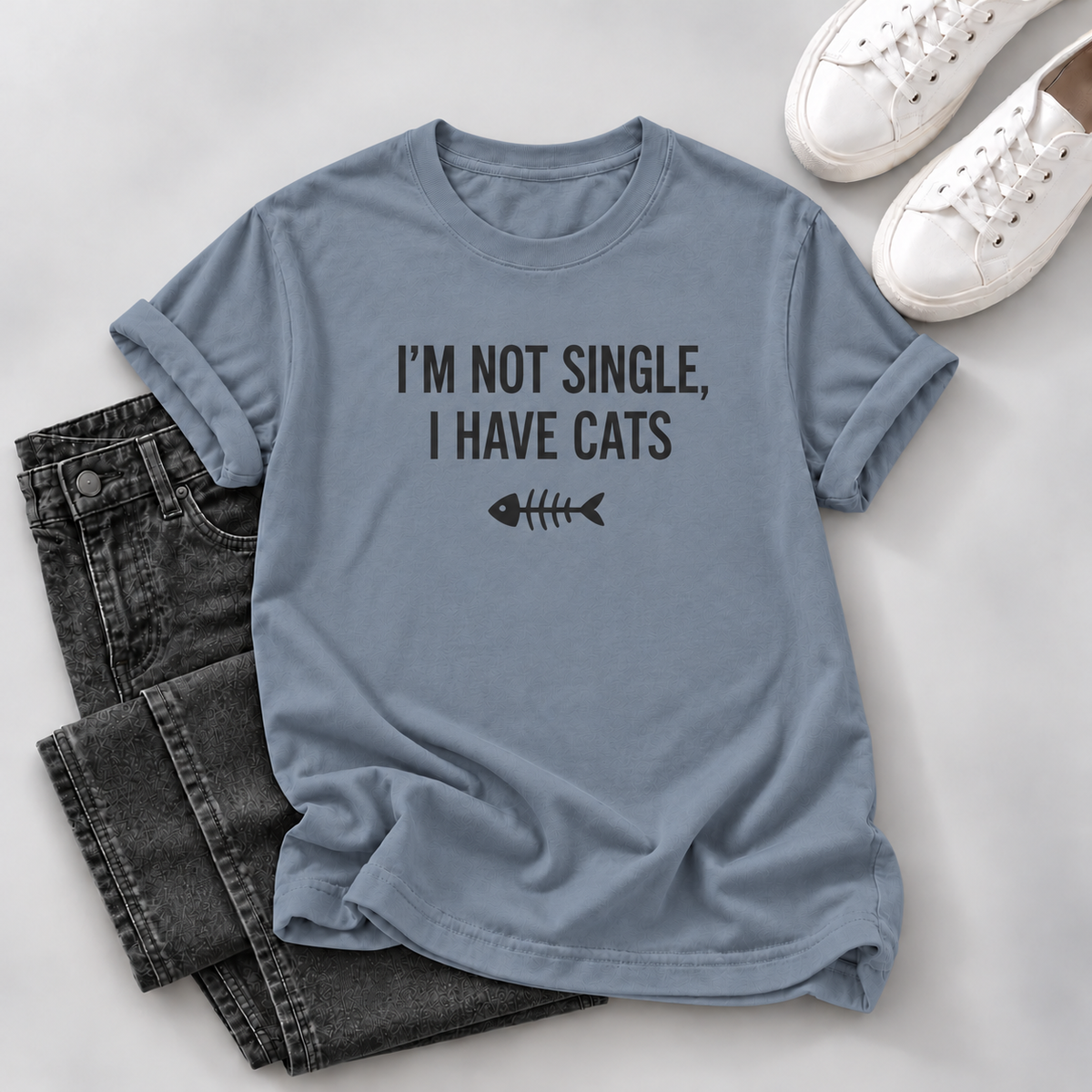 A light blue graphic t-shirt with the text 'I'M NOT SINGLE, I HAVE CATS' and a fish skeleton icon, styled in a flat lay with dark gray jeans and white sneakers on a light gray background.