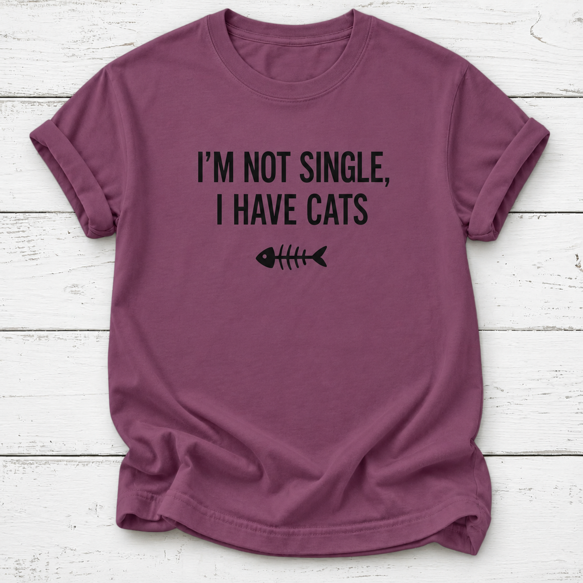 A mauve-purple short-sleeve t-shirt with black text reading 'I'M NOT SINGLE, I HAVE CATS' and a fish skeleton graphic, laid flat on a white wood plank background.