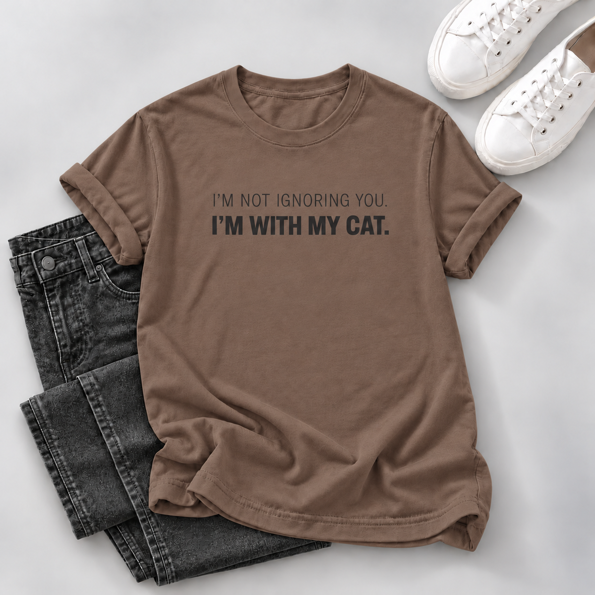 A brown short-sleeve graphic T-shirt laid flat on a light gray surface, styled with folded dark gray jeans and white sneakers. The shirt has black printed text reading, 'I'M NOT IGNORING YOU.