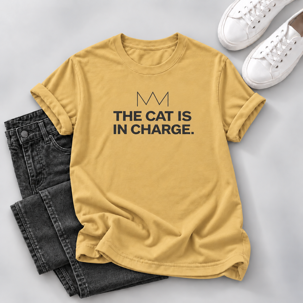 Mustard yellow outfit: Mustard yellow T-shirt with black “The Cat Is in Charge” lettering and a crown graphic, styled with dark jeans and white sneakers on a gray background.