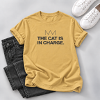 Mustard yellow outfit: Mustard yellow T-shirt with black “The Cat Is in Charge” lettering and a crown graphic, styled with dark jeans and white sneakers on a gray background.