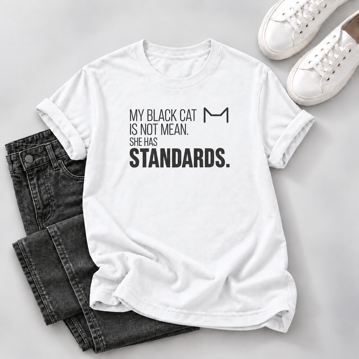 White T-shirt with black “My Black Cat Is Not Mean. She Has Standards.” lettering and a cat-ear outline, styled with dark jeans and white sneakers.