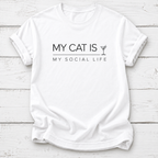 A white short-sleeve crewneck t-shirt with black printed text reading 'MY CAT IS MY SOCIAL LIFE' and a small martini glass icon, laid flat on a white wood plank background.
