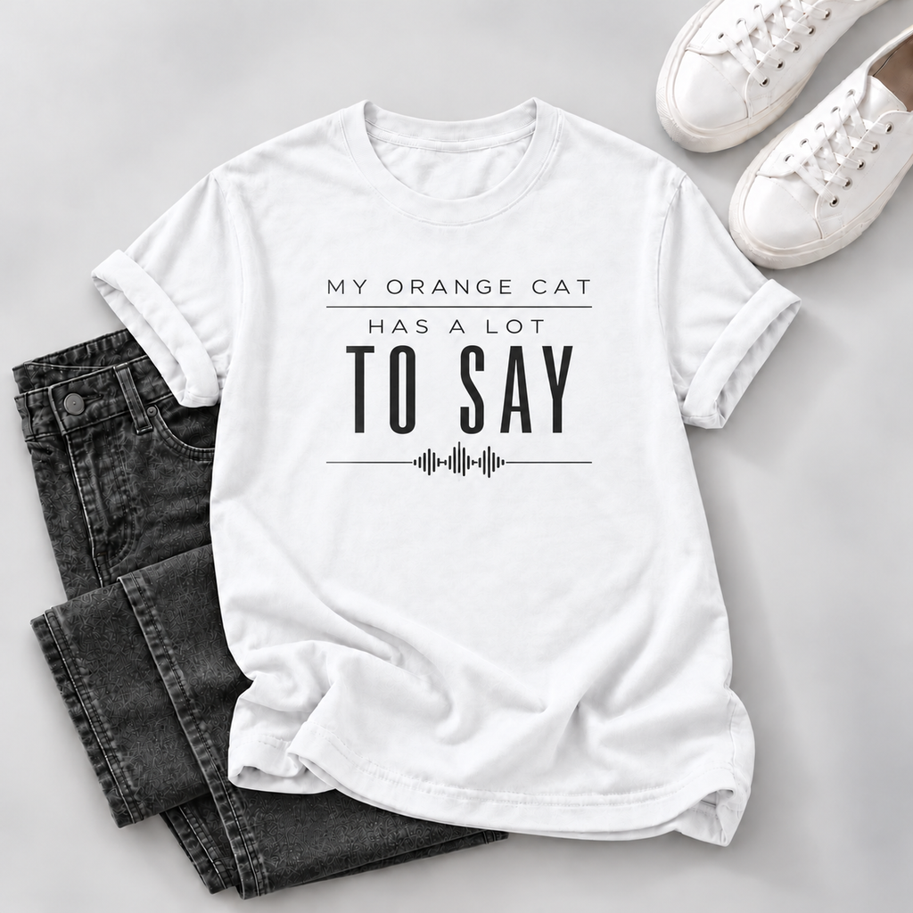 A white short-sleeve graphic T-shirt laid flat on a light gray surface, styled with folded dark gray jeans and white sneakers. The shirt has black text reading 'MY ORANGE CAT HAS A LOT TO SAY' with a soundwave graphic below.