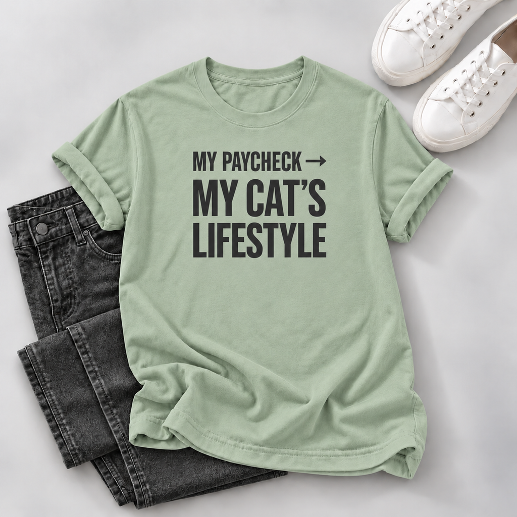 A sage green graphic t-shirt with the text 'MY PAYCHECK -> MY CAT'S LIFESTYLE' in black, styled in a flat lay with dark gray jeans and white sneakers on a light gray background.