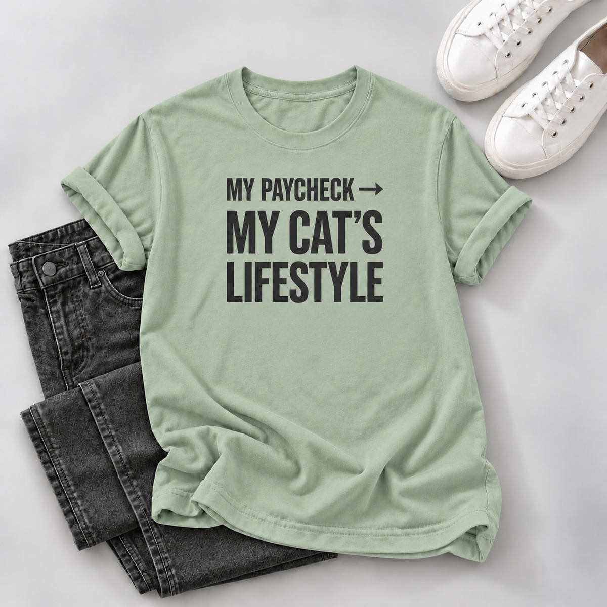 A sage green graphic t-shirt with the text 'MY PAYCHECK -> MY CAT'S LIFESTYLE' in black, styled in a flat lay with dark gray jeans and white sneakers on a light gray background.
