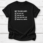 A black short-sleeve t-shirt with a white printed graphic that reads 'MY TO-DO LIST:' followed by four checked items: 'PET MY CAT', 'FEED MY CAT', 'PLAY WITH MY CAT', and 'REPEAT ALL ABOVE.' The shirt is laid flat on a white wood-plank background.
