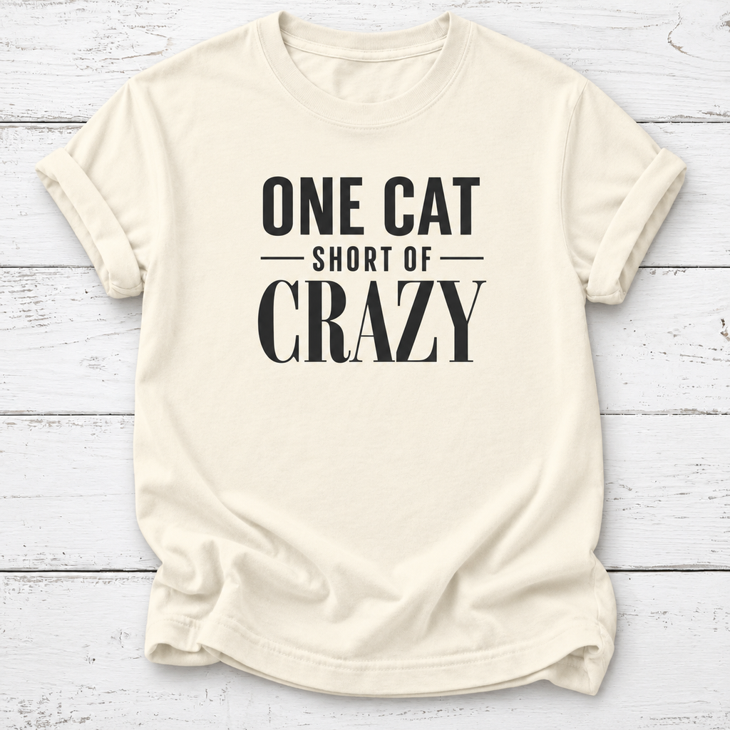 An off-white short-sleeve t-shirt with rolled sleeves, featuring the text 'ONE CAT SHORT OF CRAZY' printed in black on the front, laid flat on a white wood plank background.