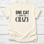 An off-white short-sleeve t-shirt with rolled sleeves, featuring the text 'ONE CAT SHORT OF CRAZY' printed in black on the front, laid flat on a white wood plank background.