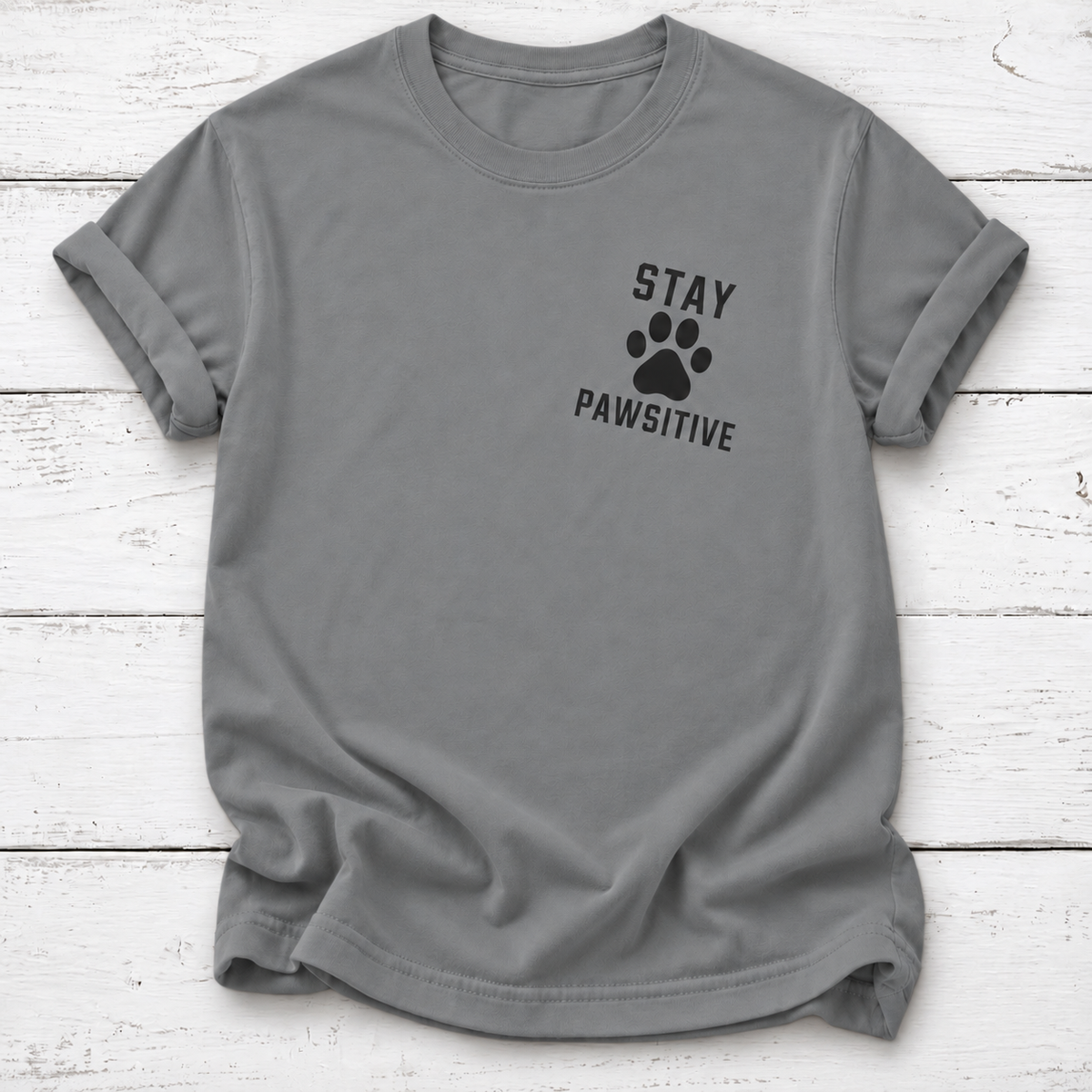 Gray T-shirt with a black paw print between “Stay” and “Pawsitive” on the left chest, with rolled sleeves, laid flat on white wooden board