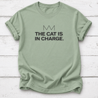 Sage green tee: Sage green T-shirt with black “The Cat Is in Charge” lettering and a minimalist crown graphic, laid flat on white wooden boards.