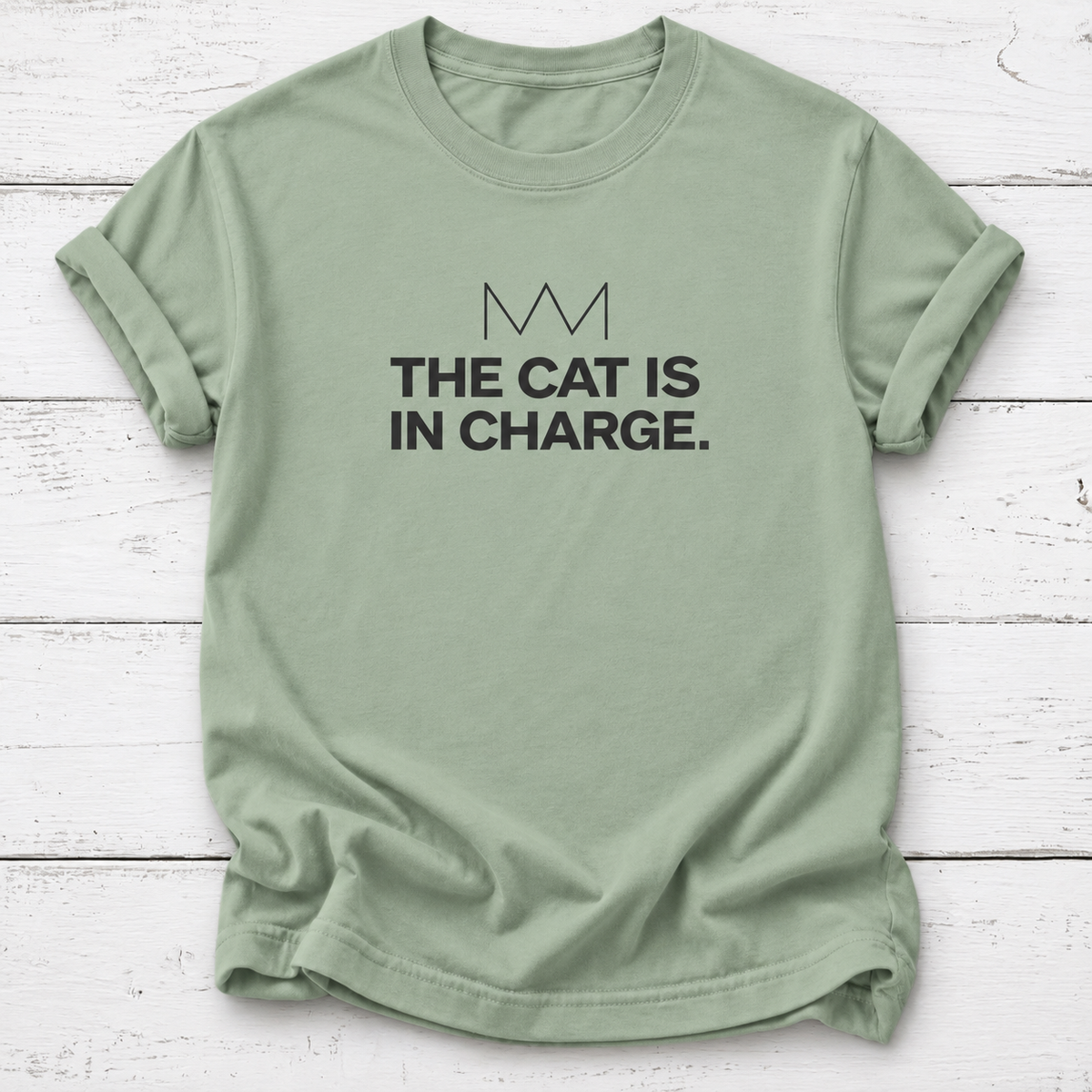 Sage green tee: Sage green T-shirt with black “The Cat Is in Charge” lettering and a minimalist crown graphic, laid flat on white wooden boards.